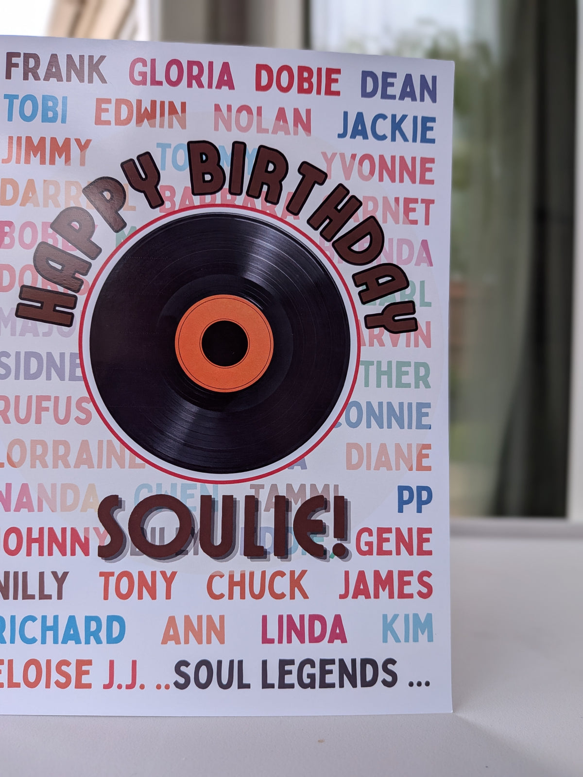A birthday card featuring a black vinyl record graphic with an orange center and the words 'HAPPY BIRTHDAY SOULIE!' in bold lettering. The card background is white with a collage of colorful song names, and the photo is taken on a light surface with a softly blurred indoor background.
