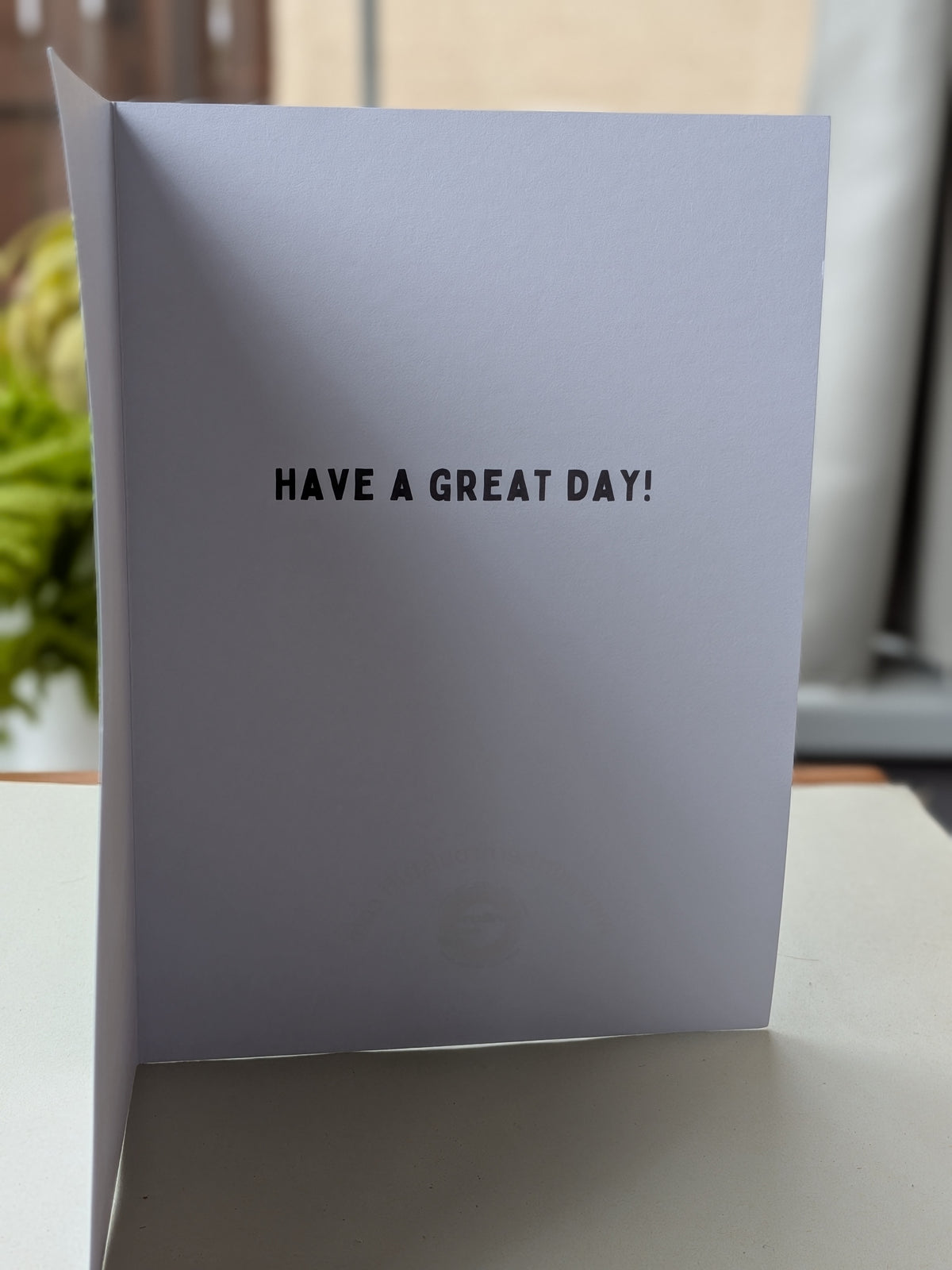 A white greeting card inner standing upright on a tabletop, printed with the phrase 'HAVE A GREAT DAY!' in black uppercase letters. The background is softly blurred with indoor lighting.