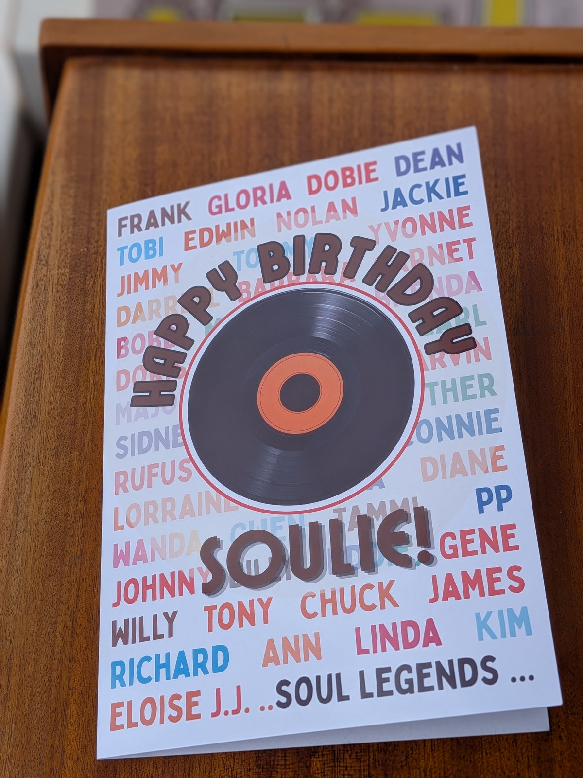 A 'Soulie' birthday card featuring a vinyl record graphic, displayed on a wooden surface. The card has a white background with a colorful array of names and the text 'HAPPY BIRTHDAY SOULIE!'.