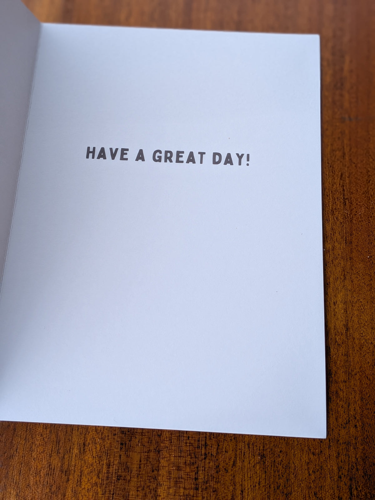 A white greeting card inner laid on a brown table, printed with the phrase 'HAVE A GREAT DAY!' in black uppercase letters. 