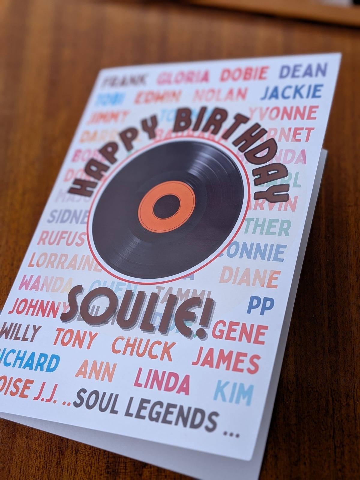 Soulie' birthday card featuring a vinyl record graphic,laid on a brown table surface. The card has a white background with a colorful array of names and the text 'HAPPY BIRTHDAY SOULIE!'.