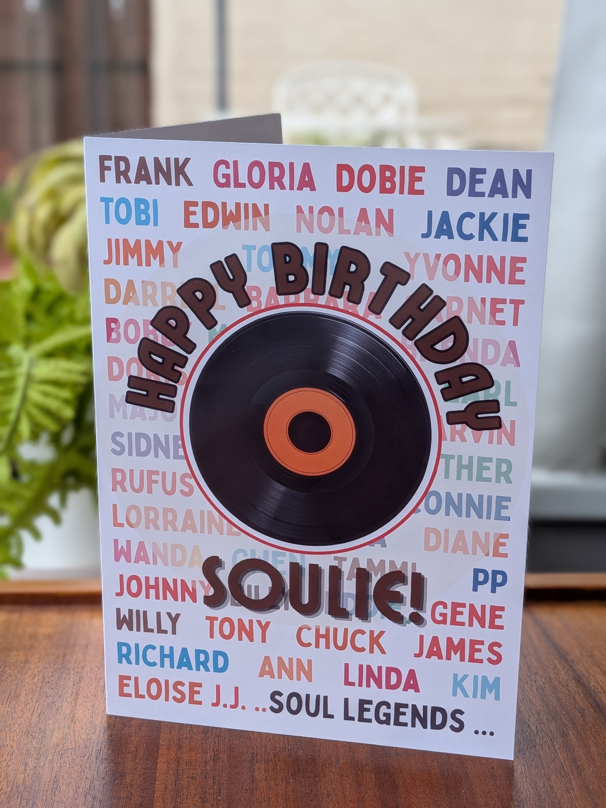 A 'Happy Birthday Soulie greeting card featuring a black vinyl record graphic with an orange center, set on a wooden table with a blurred indoor background.