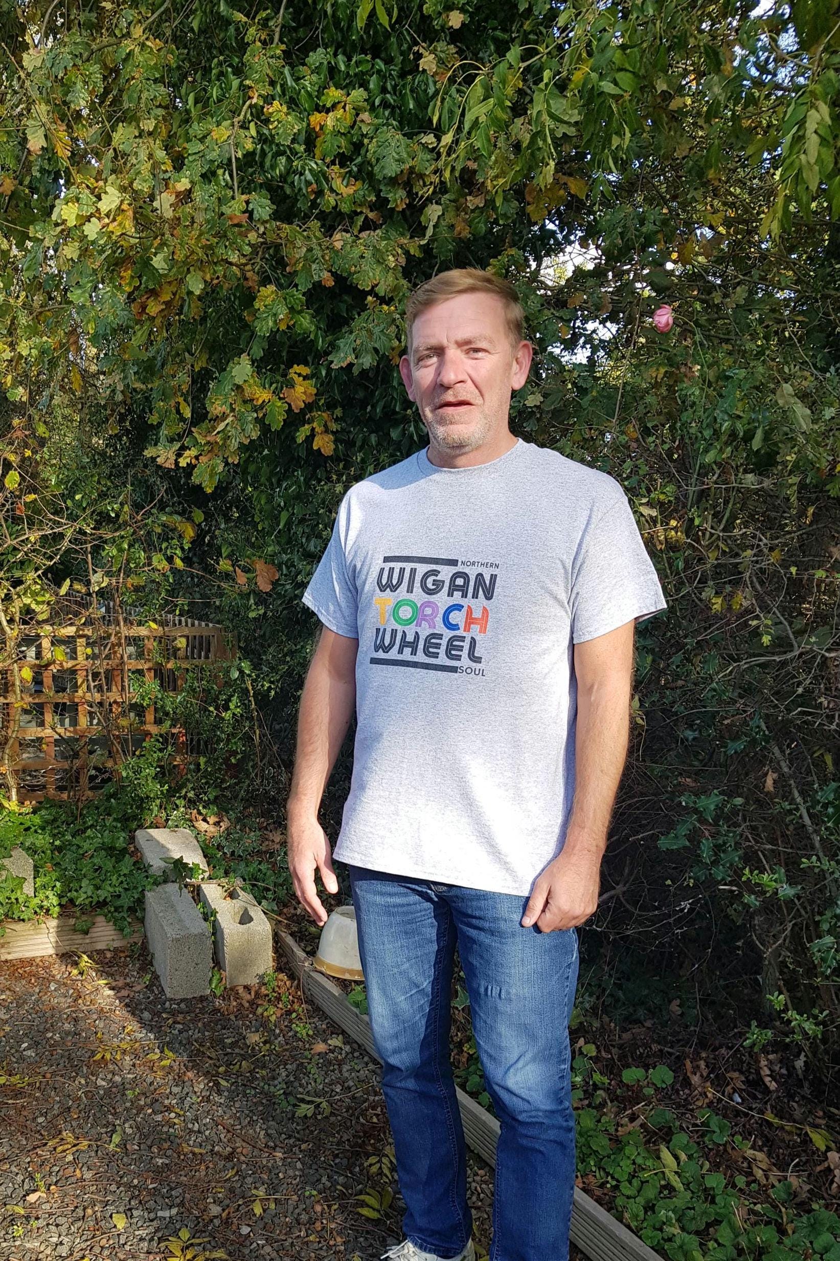 Man wearing a 'Wigan Torch Wheel' t-shirt standing outdoors with greenery in the background