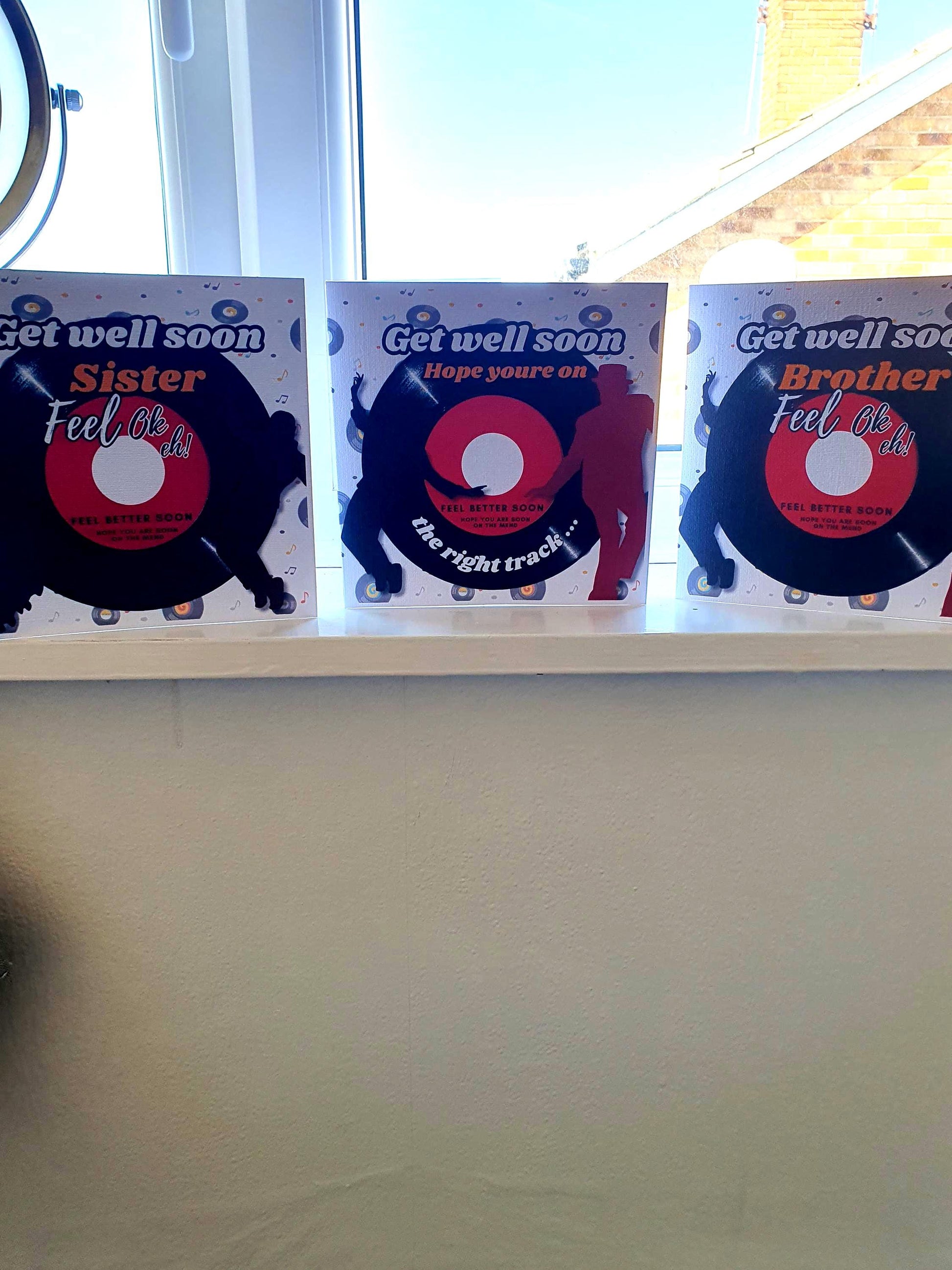 Three 'Get well soon' cards on a shelf with a window in the background. One states SISTER one states BROTHER and states ON THE RIGHT TRACK